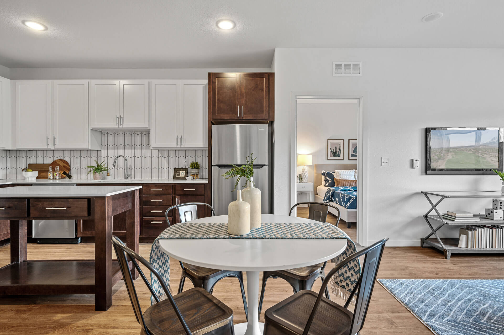 Modern kitchen with white cabinetry, stainless steel appliances, and a contrasting dark wood island, leading to a dining area with a round white table, patterned chairs, and a view into a cozy bedroom with visible decor.