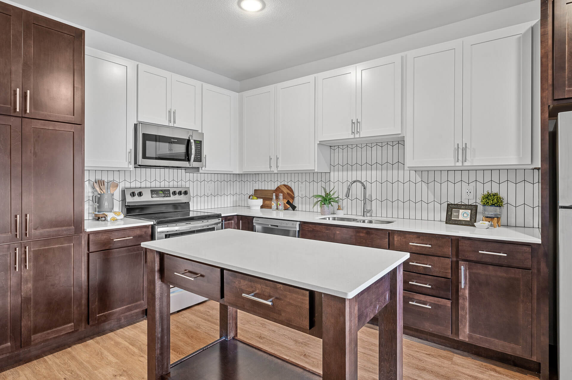 Contemporary kitchen with dark wood cabinetry below white countertops, white upper cabinets, and modern stainless steel appliances. The backsplash is a white tile with black grout, and there's a central island for additional workspace and seating.