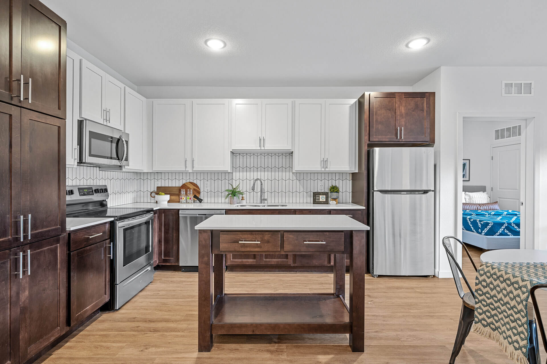 Bright kitchen with a mix of white and dark wood cabinets, stainless steel appliances, and a central kitchen island. The open layout reveals a glimpse into a dining area and a bedroom with blue bedding in the background.