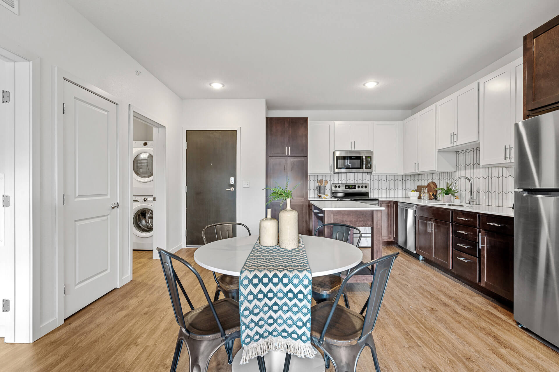 Open-plan apartment interior showing a dining area with a white round table and metal chairs, adjacent to a kitchen with white upper cabinets, dark brown lower cabinets, stainless steel appliances, and a laundry closet with a washer and dryer.