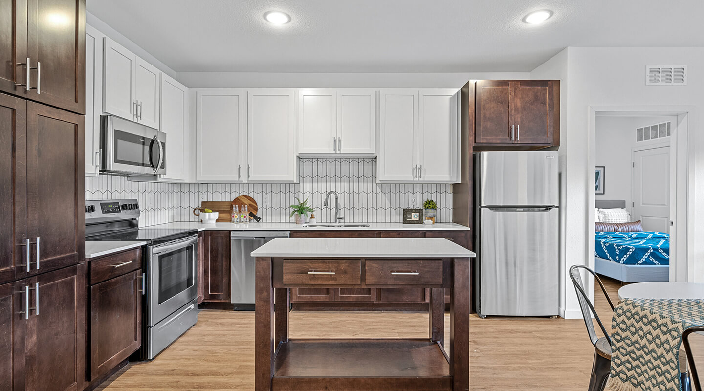 Modern kitchen with white cabinetry and stainless steel appliances, featuring a center island with dark wood cabinets, an open doorway to a bedroom, and a dining area with a patterned chair.